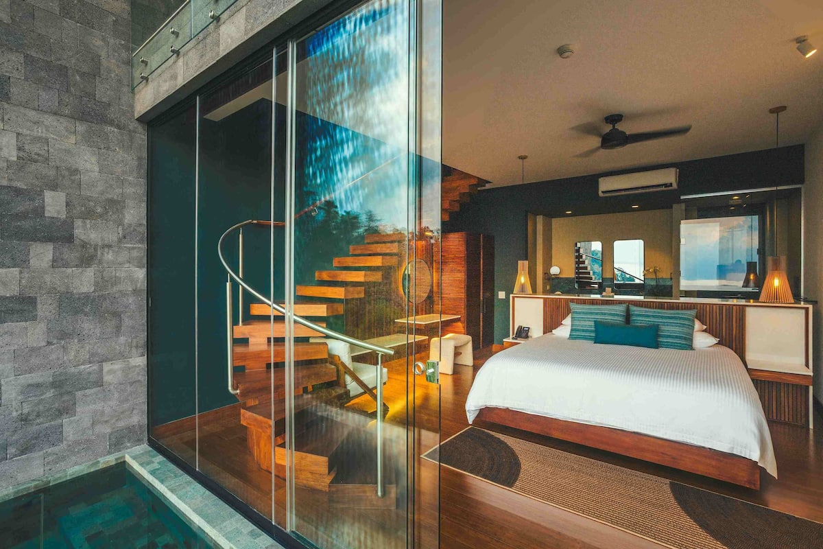 Une chambre à coucher moderne avec un grand lit, un mur en verre, un escalier en bois et une vue sur l’extérieur.
