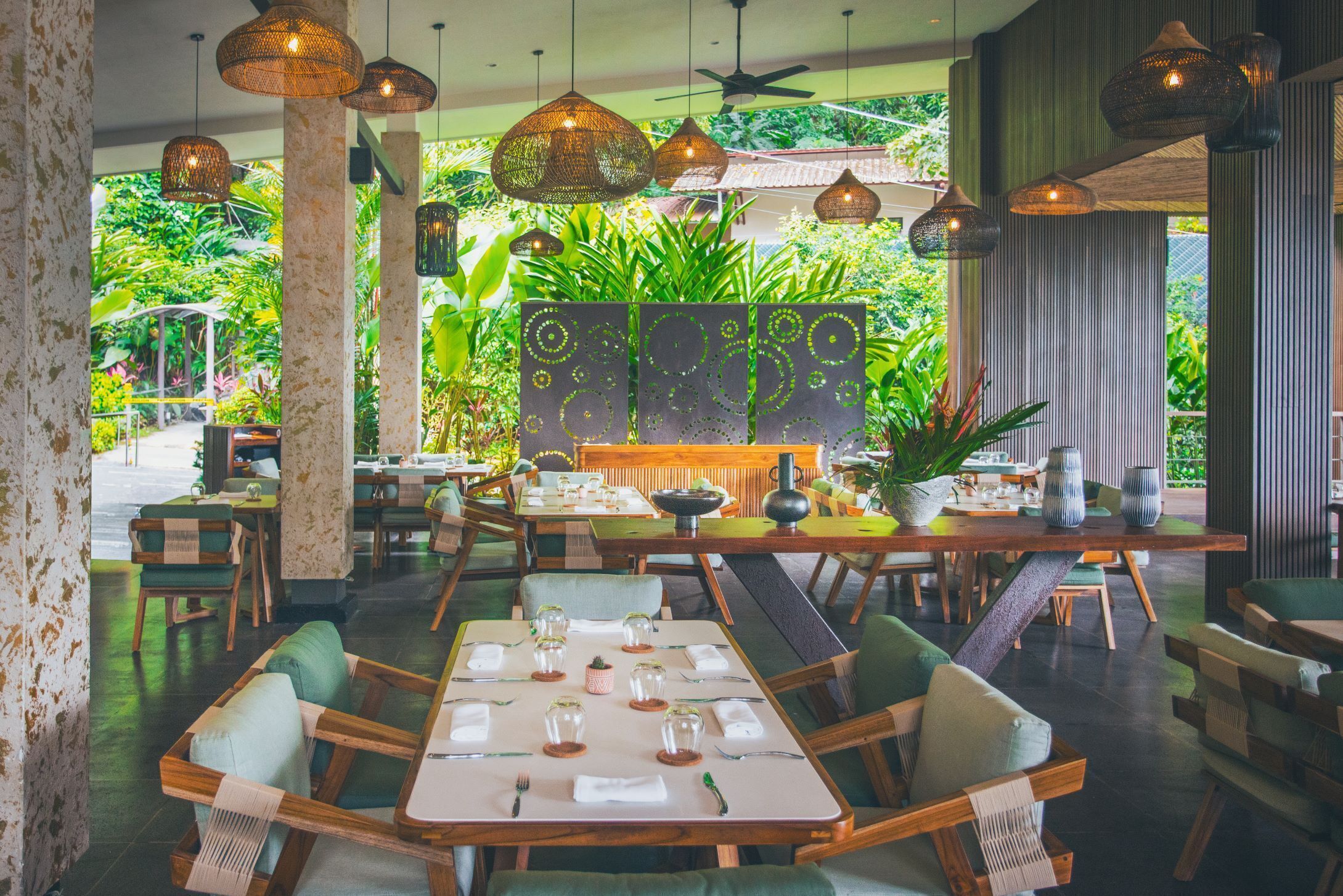 A restaurant with wooden tables and chairs, hanging pendant lights, and a view of lush greenery outside.