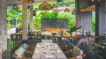 A restaurant with wooden tables and chairs, hanging pendant lights, and a view of lush greenery outside.