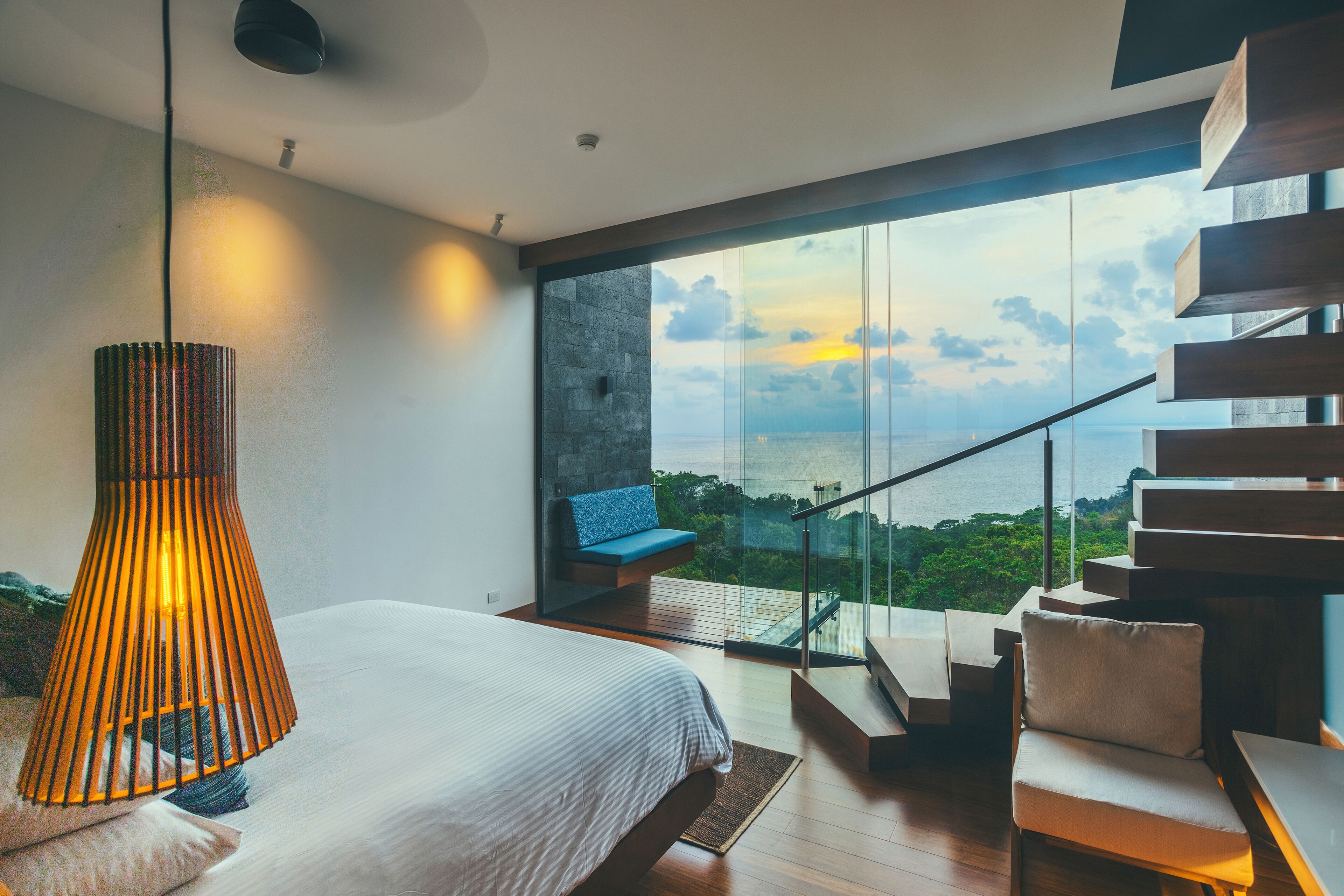 A modern bedroom with a large bed, a hanging lamp, a staircase, and a view of the sea.