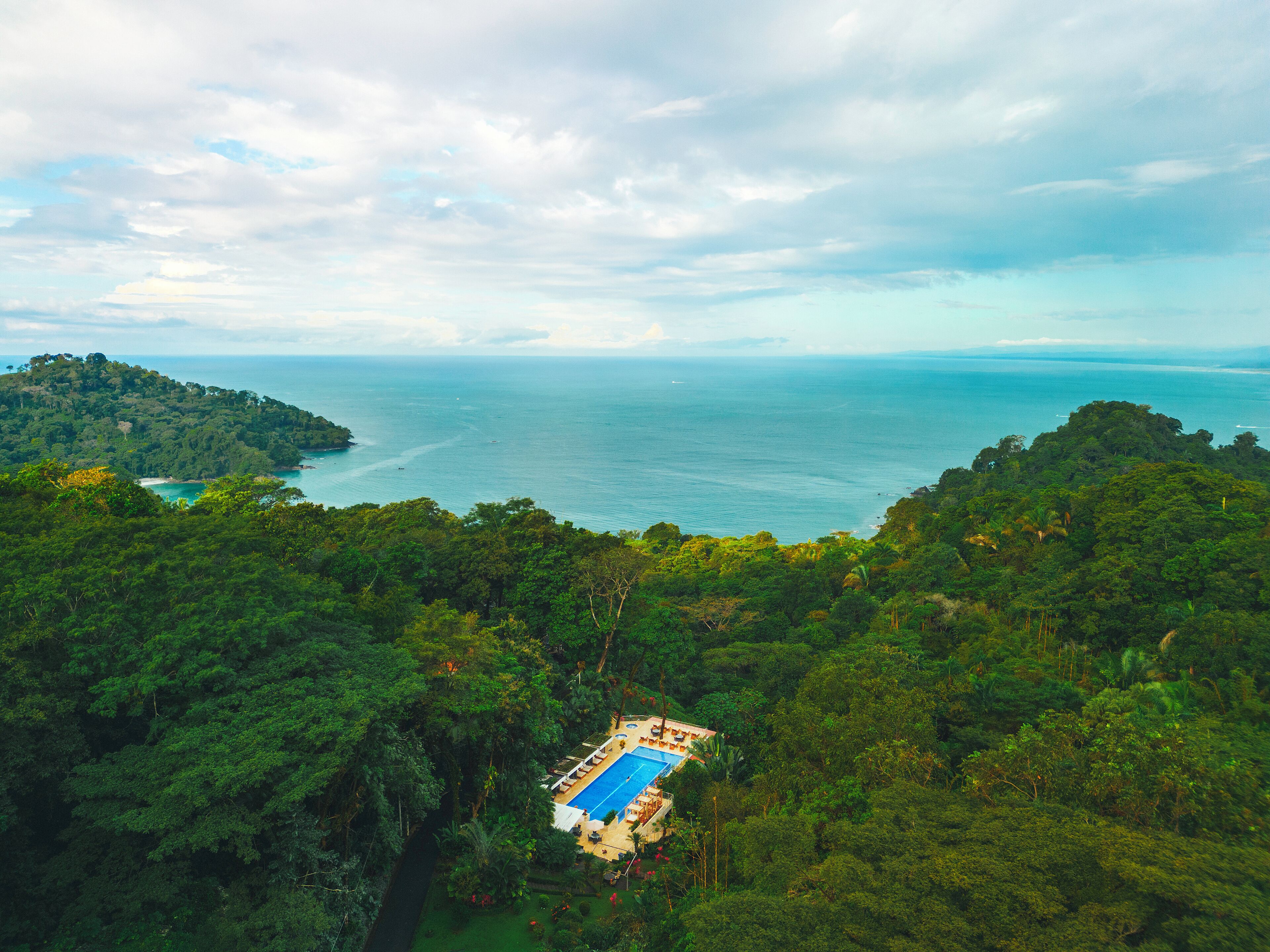 An aerial view of a tropical resort with a swimming pool surrounded by dense forest, overlooking a coastline.