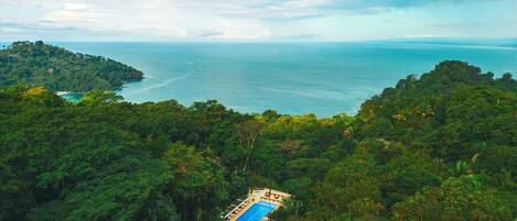 An aerial view of a tropical resort with a swimming pool surrounded by dense forest, overlooking a coastline.