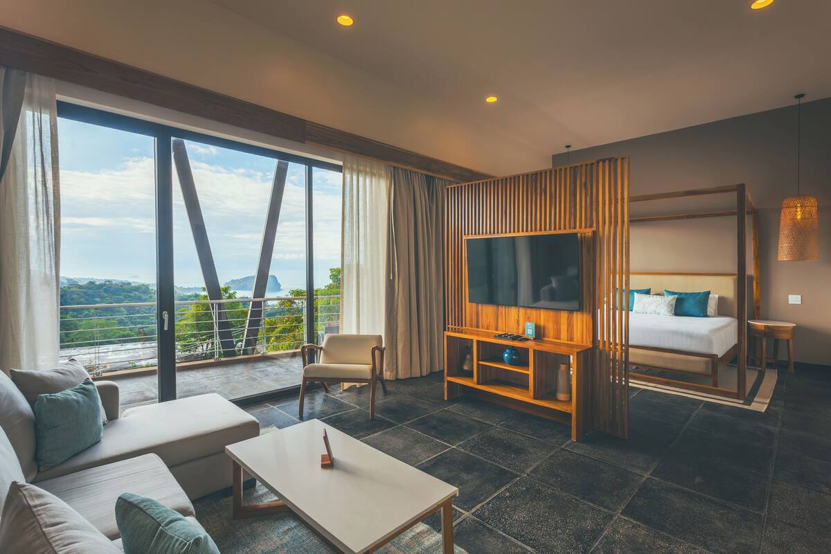 Une chambre d’hôtel moderne avec une grande fenêtre, un canapé, une télévision, un lit et une petite table.