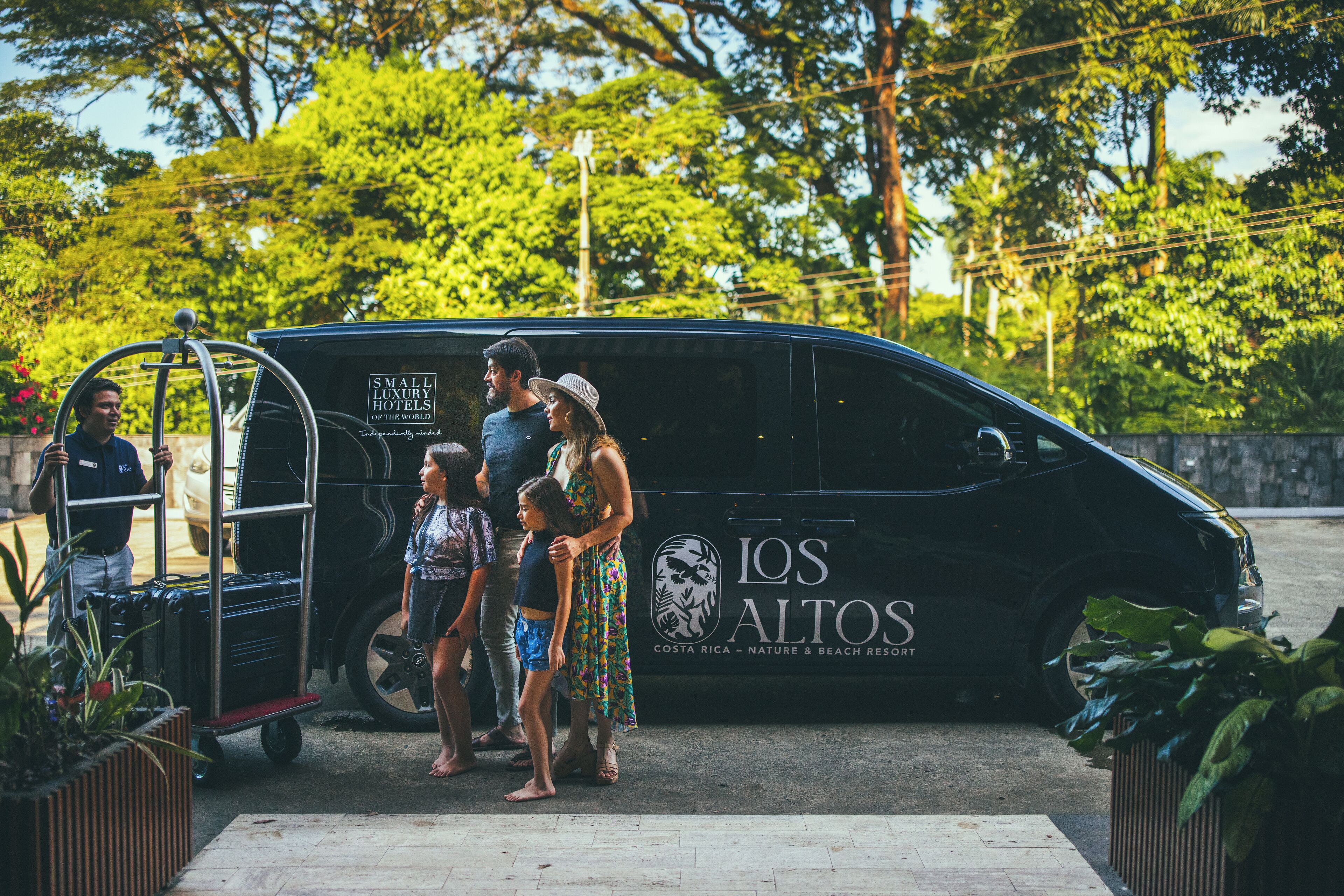 A black van with 'LOS ALTOS' and 'COSTA RICA - NATURE & BEACH RESORT' text, a luggage cart, and people standing in front.