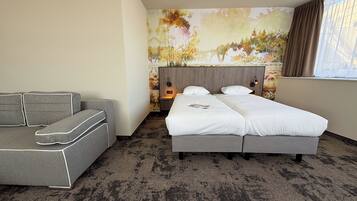 A hotel room with a grey sofa, a double bed, a nightstand with a lamp, and a mural on the wall.