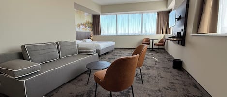 Habitación de hotel con un sofá gris, dos sillones marrones, una mesita redonda pequeña, una cama con ropa de cama blanca, un cuadro en la pared y un ventanal con cortinas.