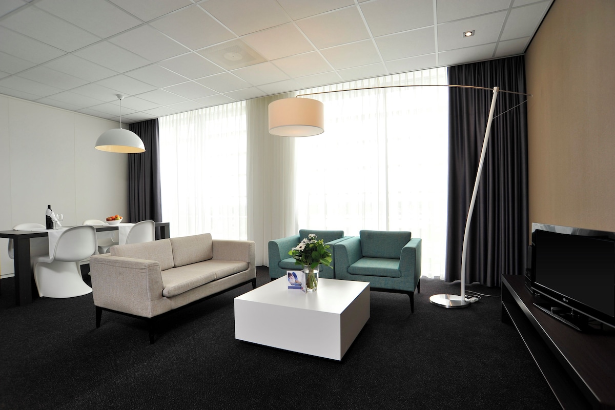 Una habitación de hotel moderna con sofá, dos sillones, una mesita de centro, televisor y un ventanal con cortinas.