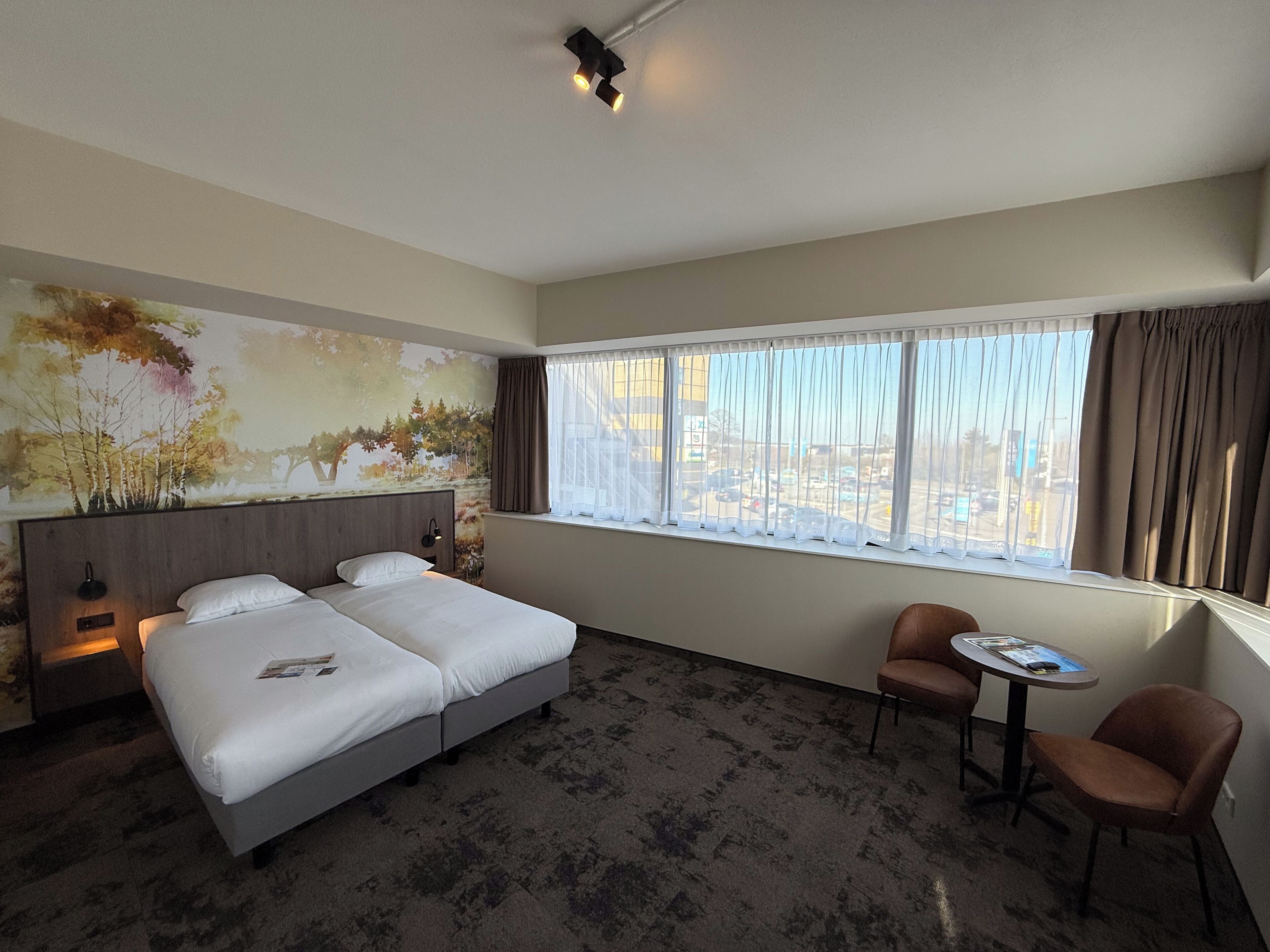 Ein Hotelzimmer mit einem großen Bett, einem Schreibtisch mit zwei Stühlen, einem Fenster mit Vorhängen und einem Wandbild.
