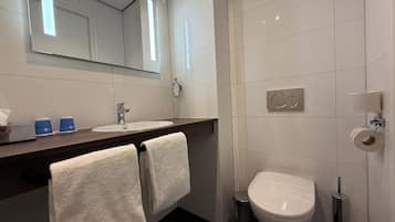 A modern bathroom with a white sink, mirror, and toilet. There are two towels hanging on a rack, a soap dispenser, and a trash can.