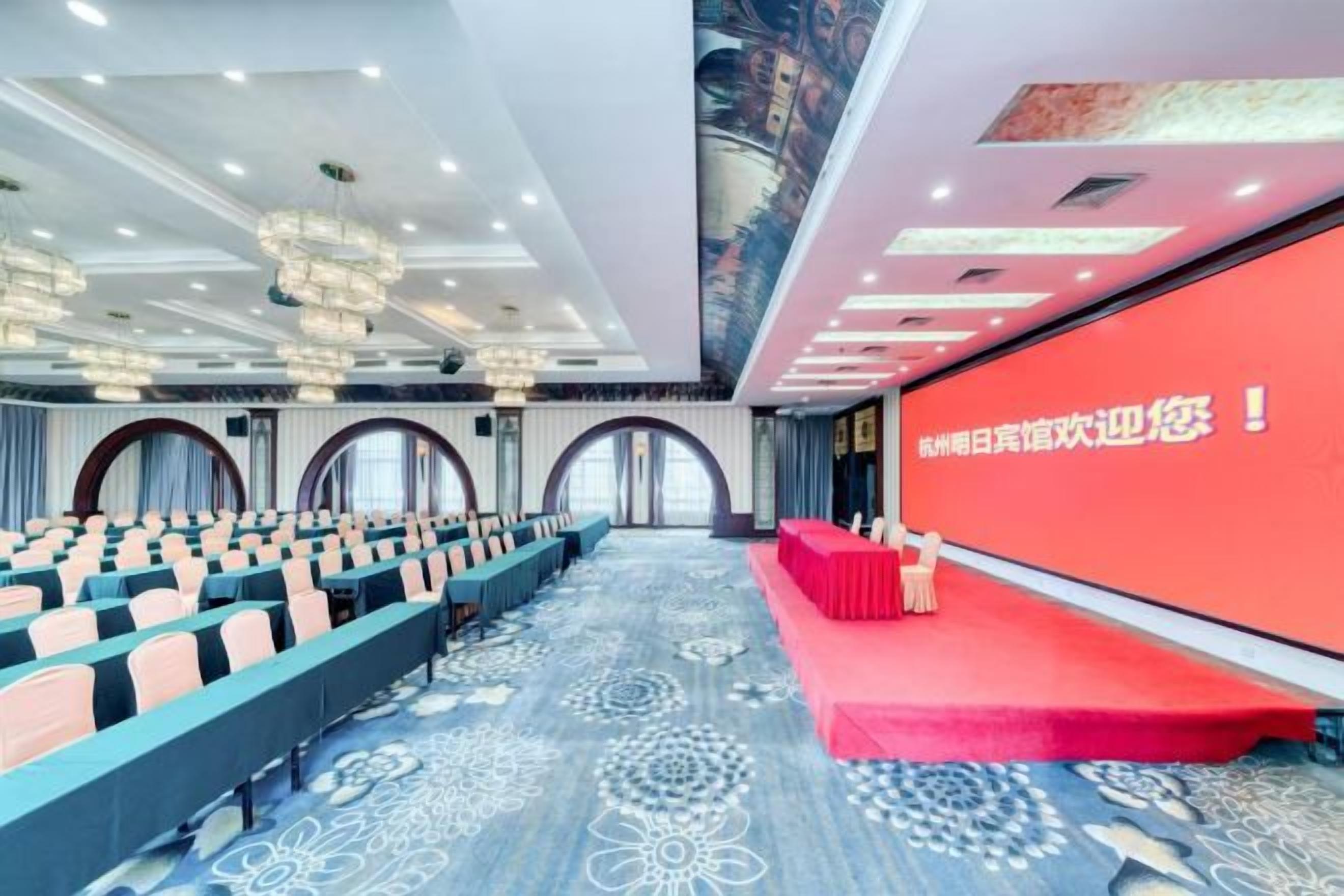 A conference room with rows of chairs, a stage with a red carpet, and a large screen with text.