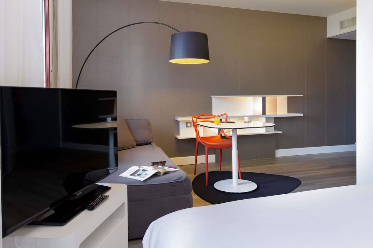 Una habitación de hotel moderna con una televisión de pantalla plana, un sofá, una pequeña mesa con una silla y una lámpara de pie grande.