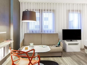 A modern hotel room with a sofa, a TV, a small table with a cup and a book, and a chair.