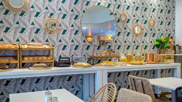 A breakfast bar with pastries and bread, surrounded by wicker chairs and a patterned wall.