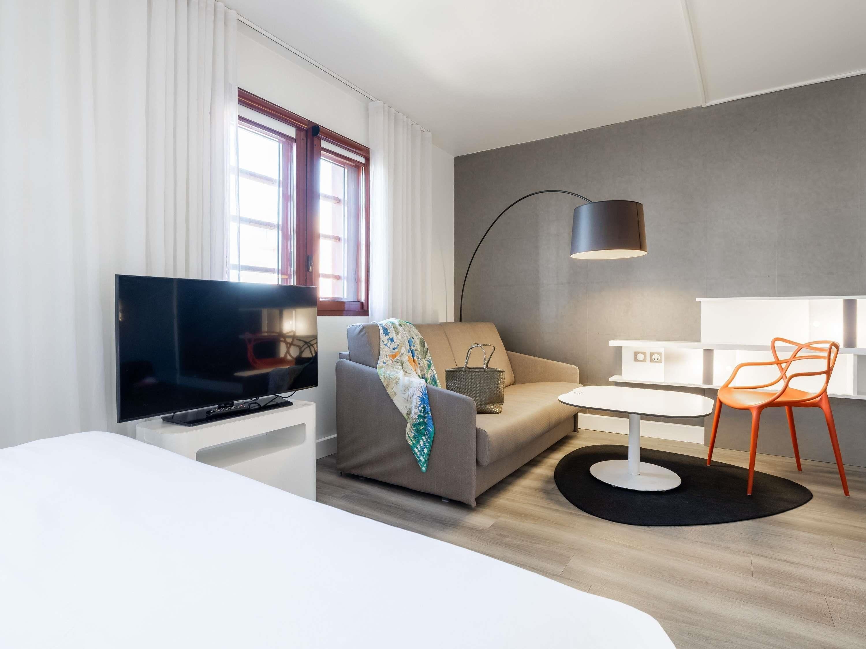 A modern hotel room with a flat-screen TV, a sofa, a small table, a lamp, and a chair.