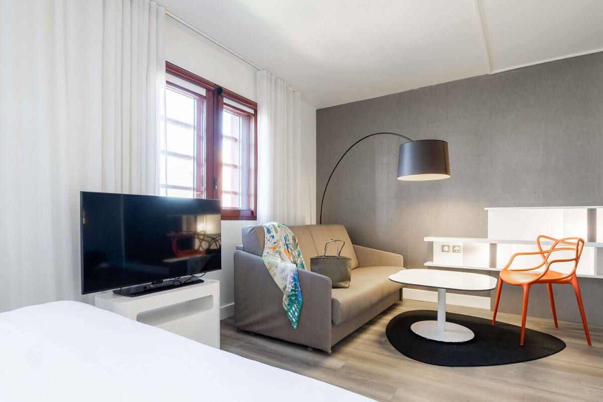 A modern hotel room with a flat-screen TV, a sofa, a small table, a lamp, and a chair.