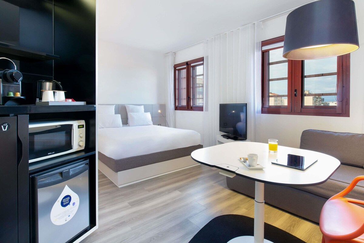 Una habitación de hotel moderna con cama, mesa de comedor, sofá, televisor, microondas y cafetera.