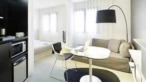 A modern hotel room with a kitchenette, a sofa, a round dining table, and a large window with blinds. - Novotel Suites Perpignan Mediterranée (Perpignan)