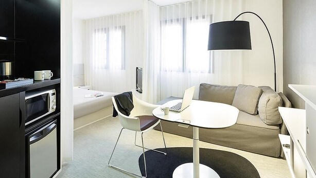 A modern hotel room with a kitchenette, a sofa, a round dining table, and a large window with blinds.