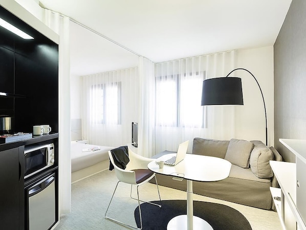 Una habitación de hotel moderna con kitchenette, un sofá, una mesa de comedor redonda y un amplio ventanal con persianas.