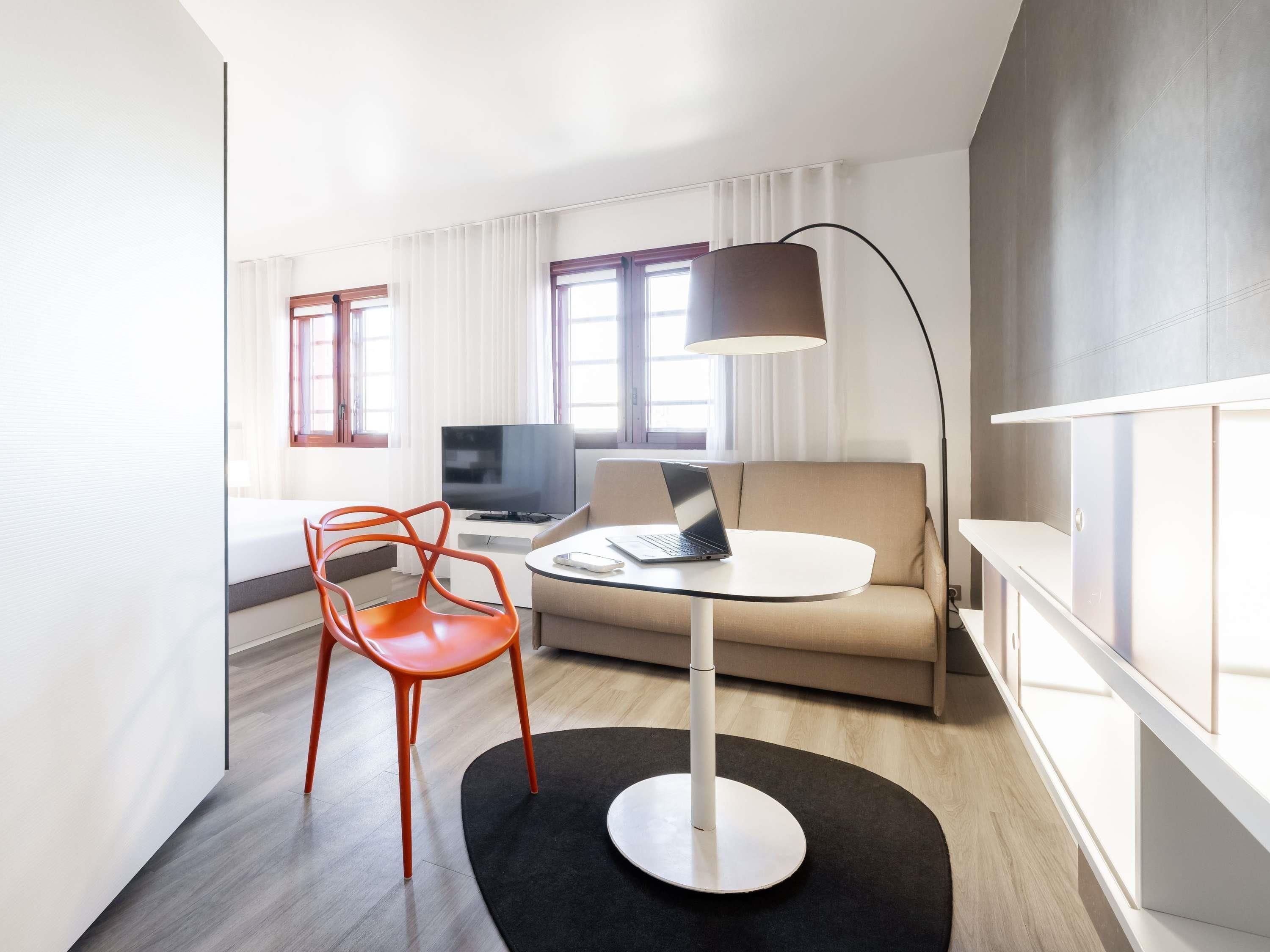 A modern hotel room with a sofa, a round table, a red chair, a TV, and a lamp.