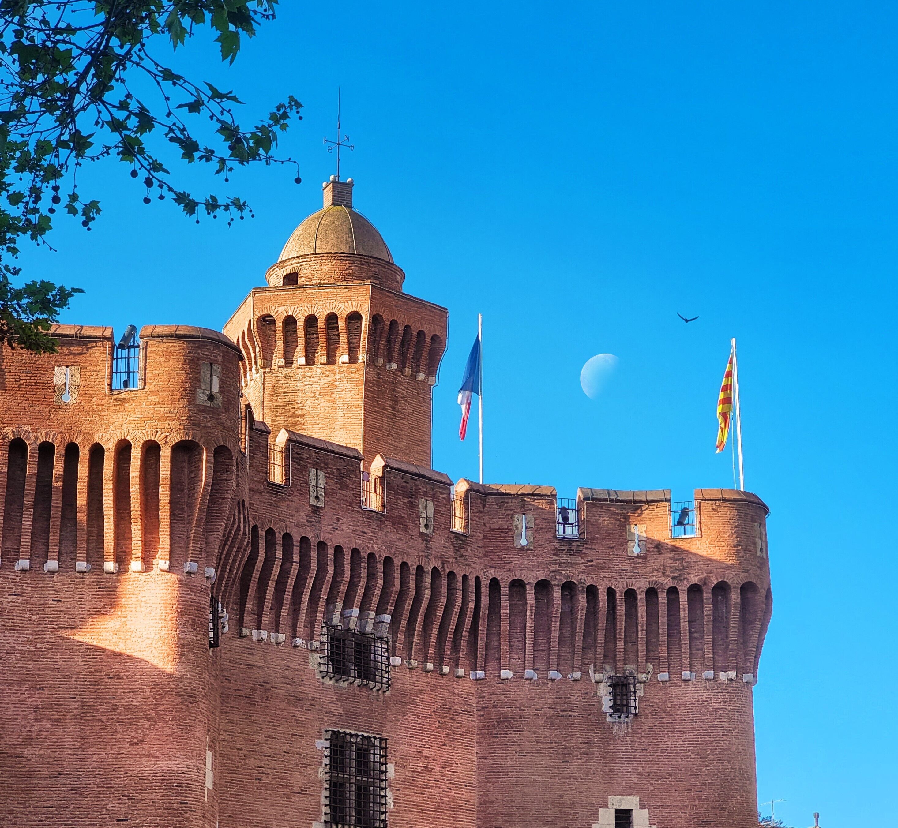 Eine historische Ziegelburg mit einem markanten Turm, mehreren Flaggen und blauem Himmel.
