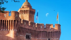 A historic brick castle with a prominent tower, multiple flags, and a clear blue sky.
