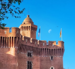 Un castillo de ladrillo histórico con una torre destacada, varias banderas y un cielo azul despejado.