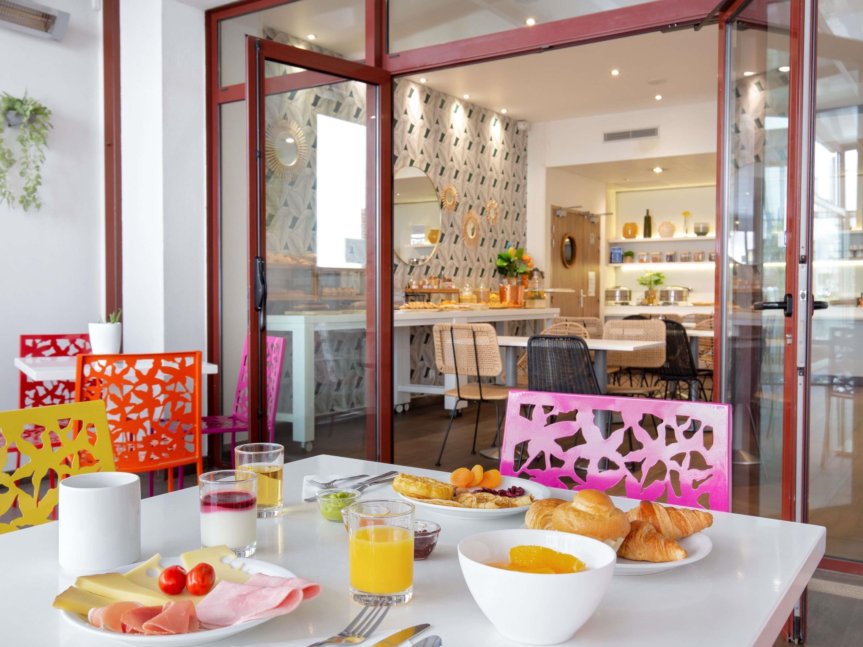 A breakfast table with various food items, a dining area with chairs and a table, and a kitchen in the background.