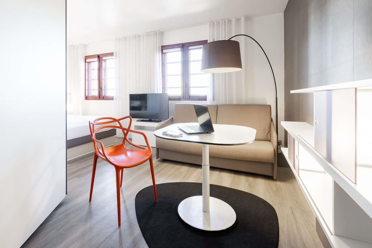 Una habitación de hotel moderna con un sofá, una mesa redonda, una computadora portátil, un televisor y una silla roja.