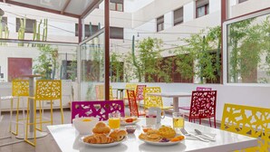 A breakfast setup with croissants, waffles, and juice on a table in a modern, open-plan dining area with yellow and pink chairs and large windows overlooking greenery. - Novotel Suites Perpignan Mediterranée (Perpignan)