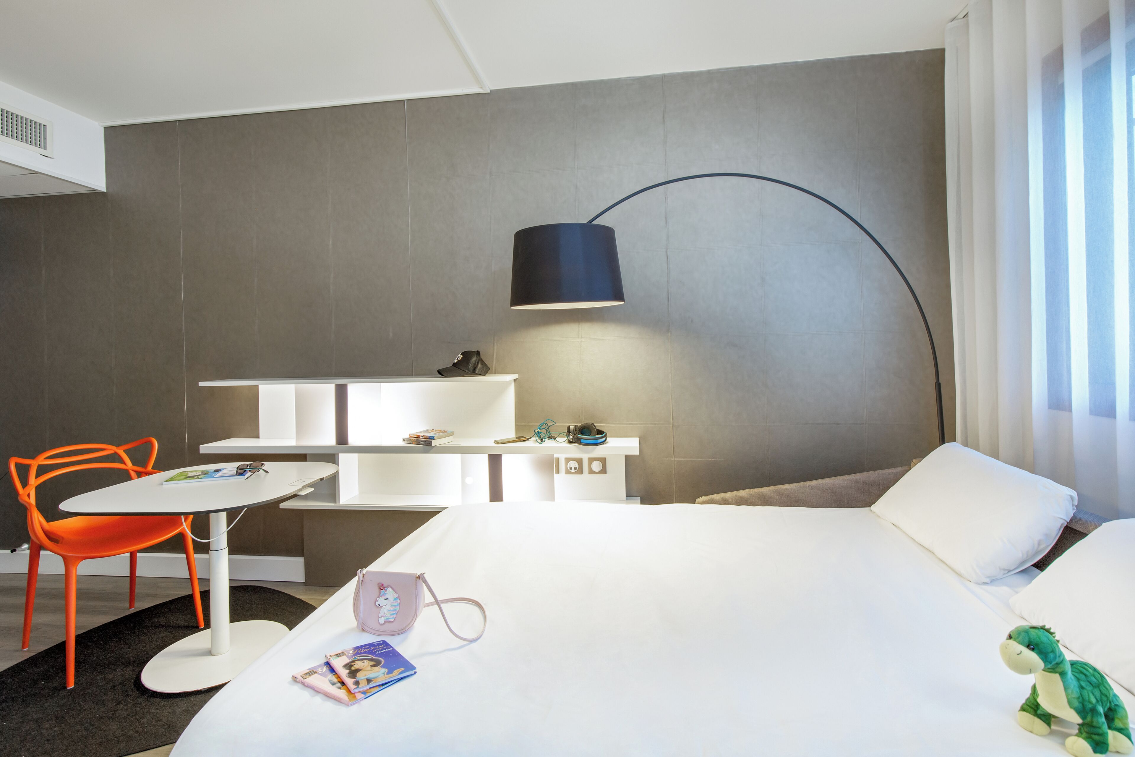 A hotel room with a bed, a desk, a chair, a lamp, and a window with curtains.
