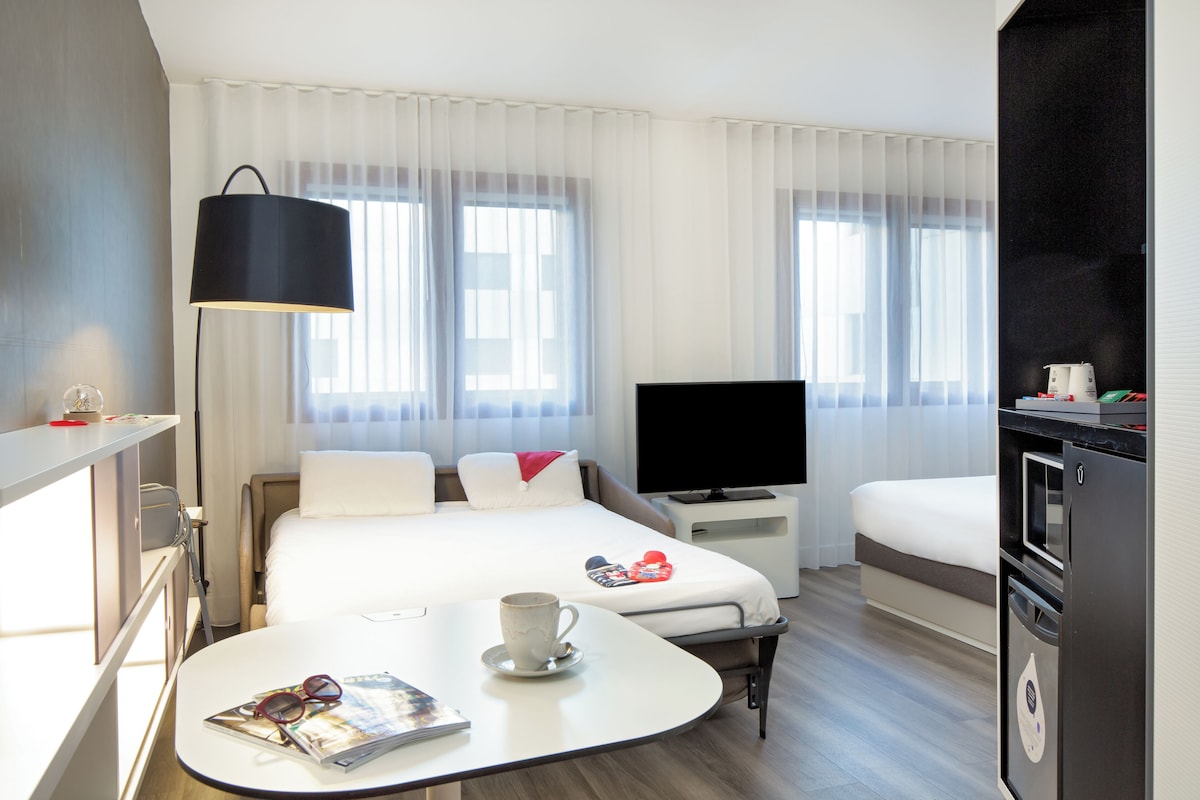 A modern hotel room with a bed, a TV, a dining table, and a kitchenette.