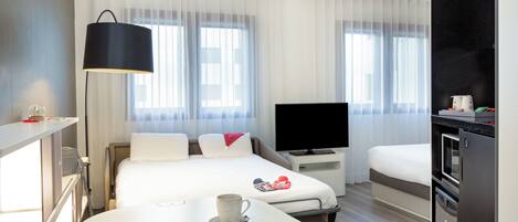A modern hotel room with a bed, a TV, a dining table, and a kitchenette.