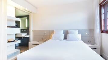 A modern hotel room with a large bed, a bedside table, a nightstand, and a built-in desk with a chair.