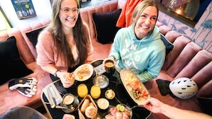 Twee personen zitten op een roze bank met een tafel vol eten en drinken voor zich.