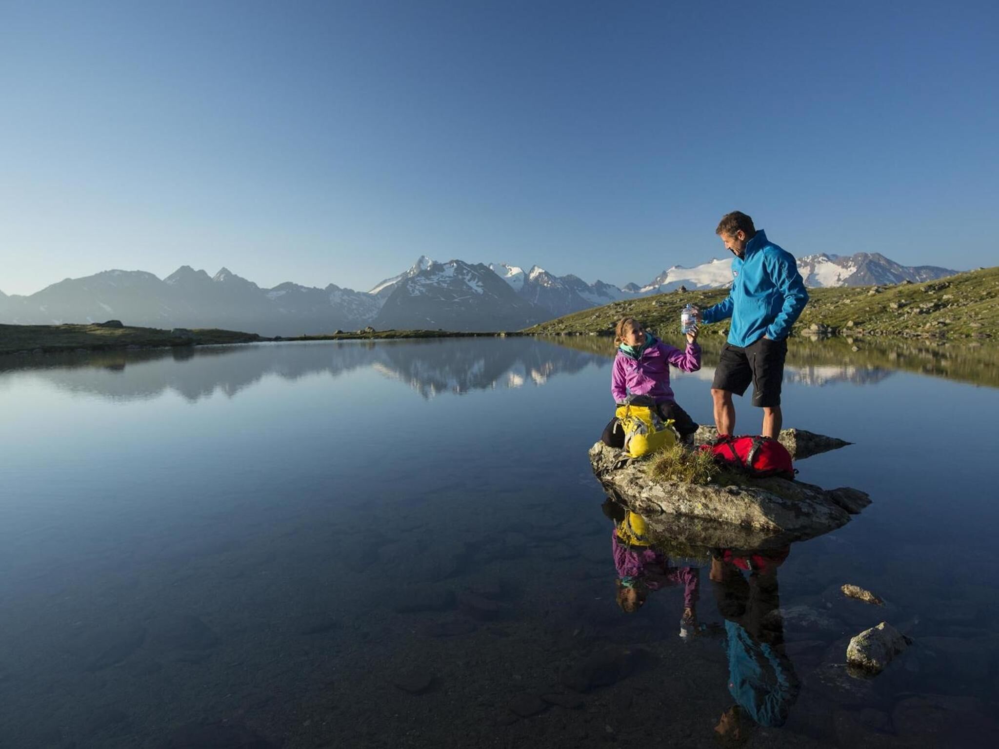 A calm lake with mountains in the background, a person standing on a rock, and another person sitting on the shore.