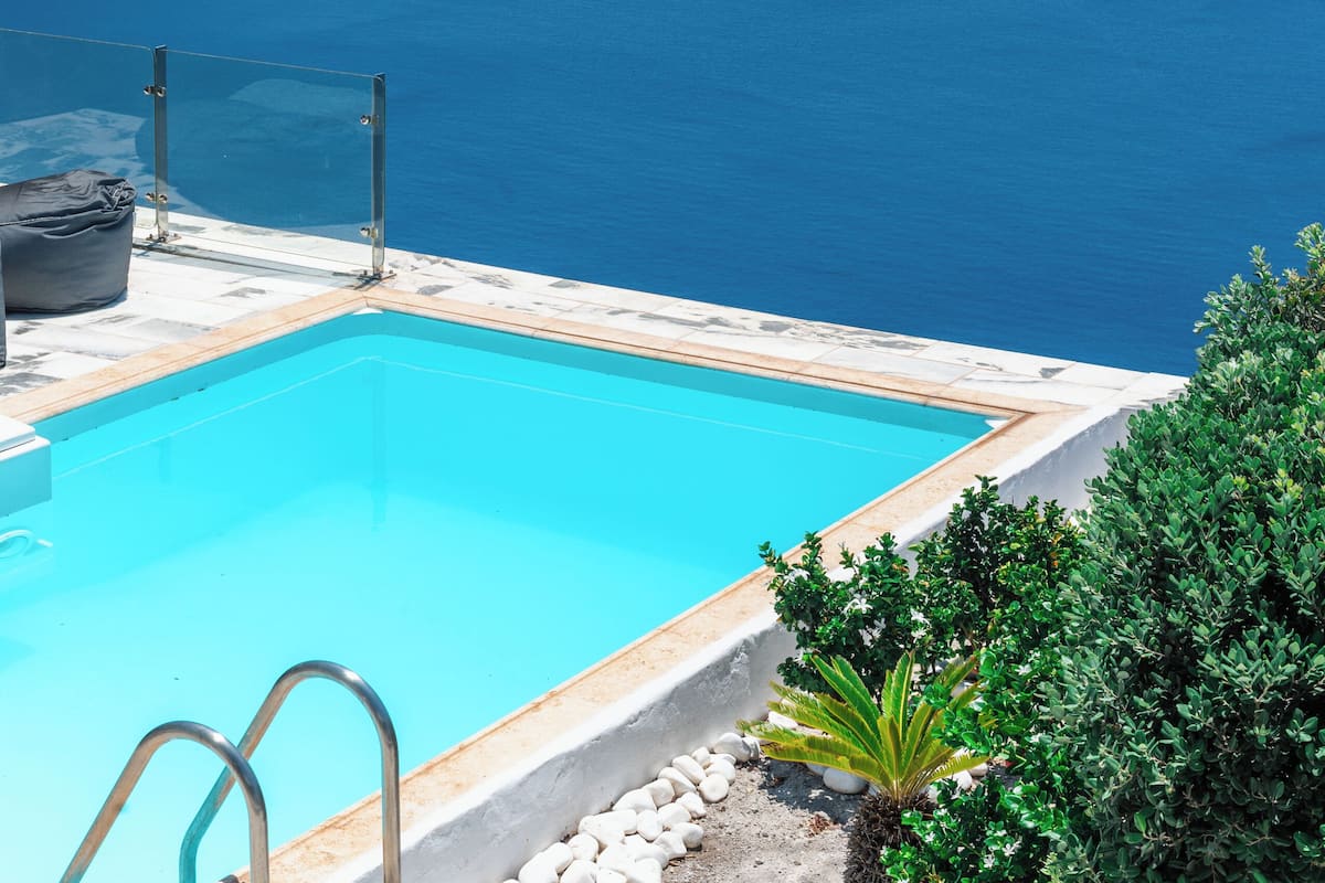 A clear blue swimming pool with a metal ladder, surrounded by white stones and greenery, overlooking a vast sea and distant islands.