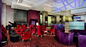 Um saguão de hotel com assentos vermelhos e roxos, um piano de cauda e uma mesa de recepção.