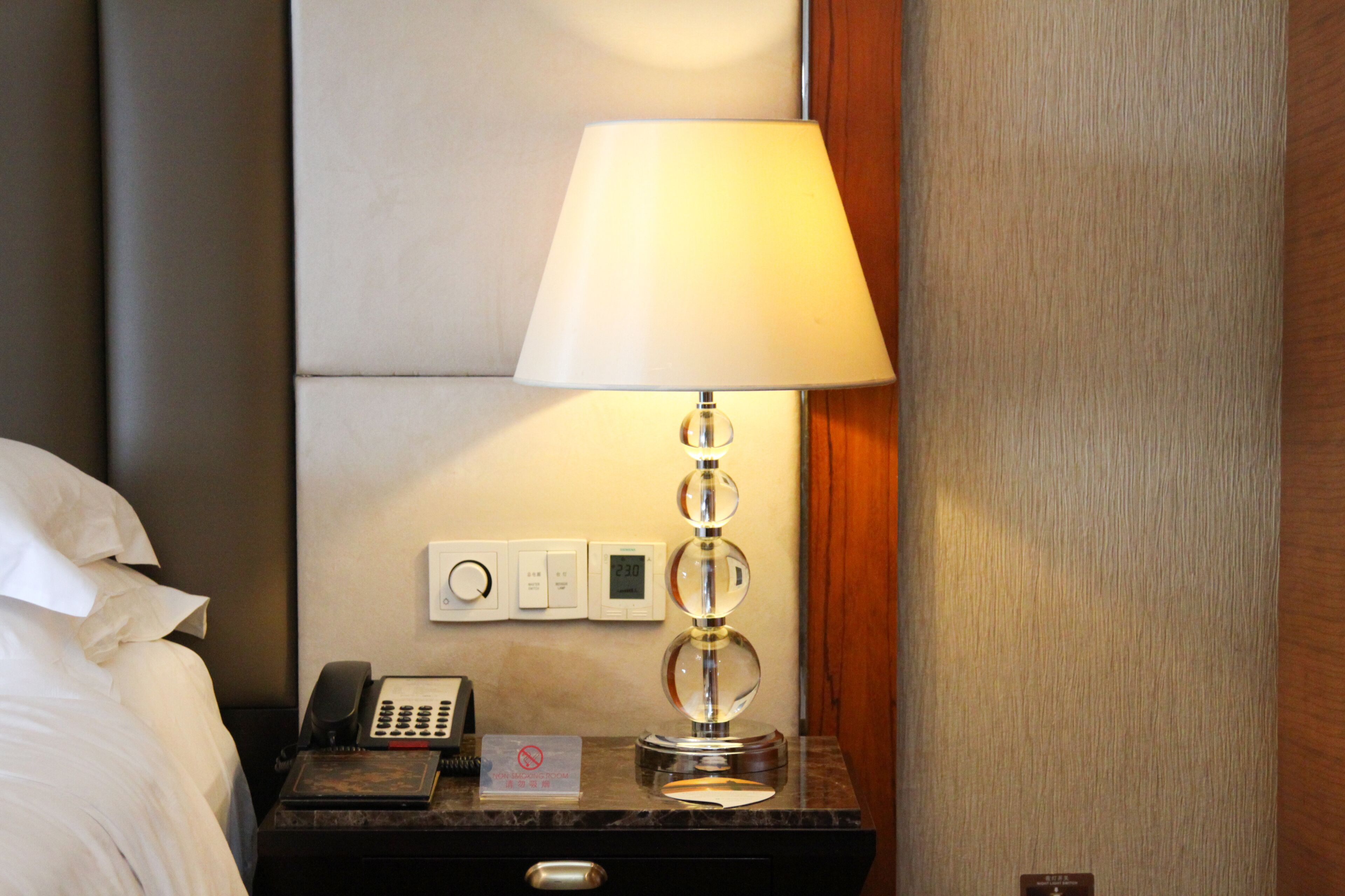 A hotel room with a bed, a nightstand with a lamp, a telephone, and a digital thermostat.