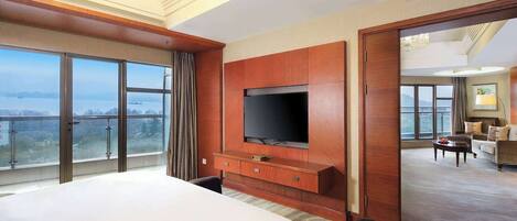 A hotel room with a large bed, a TV mounted on the wall, a balcony with a view, and a separate living area with a sofa and coffee table.