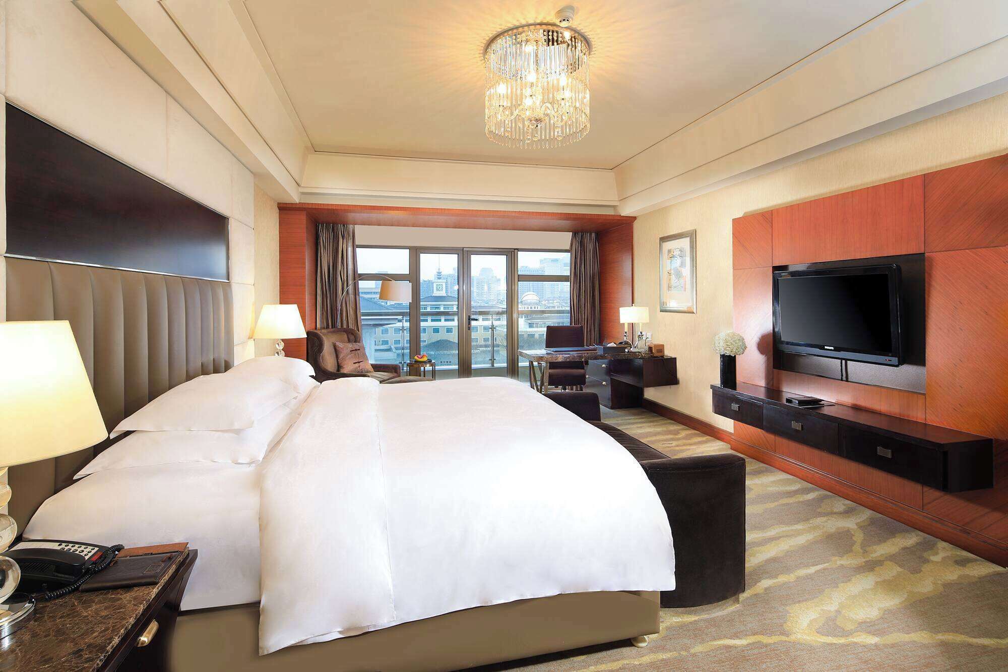 Habitación de hotel con una cama grande, televisión, escritorio y vista a la ciudad a través de las ventanas.