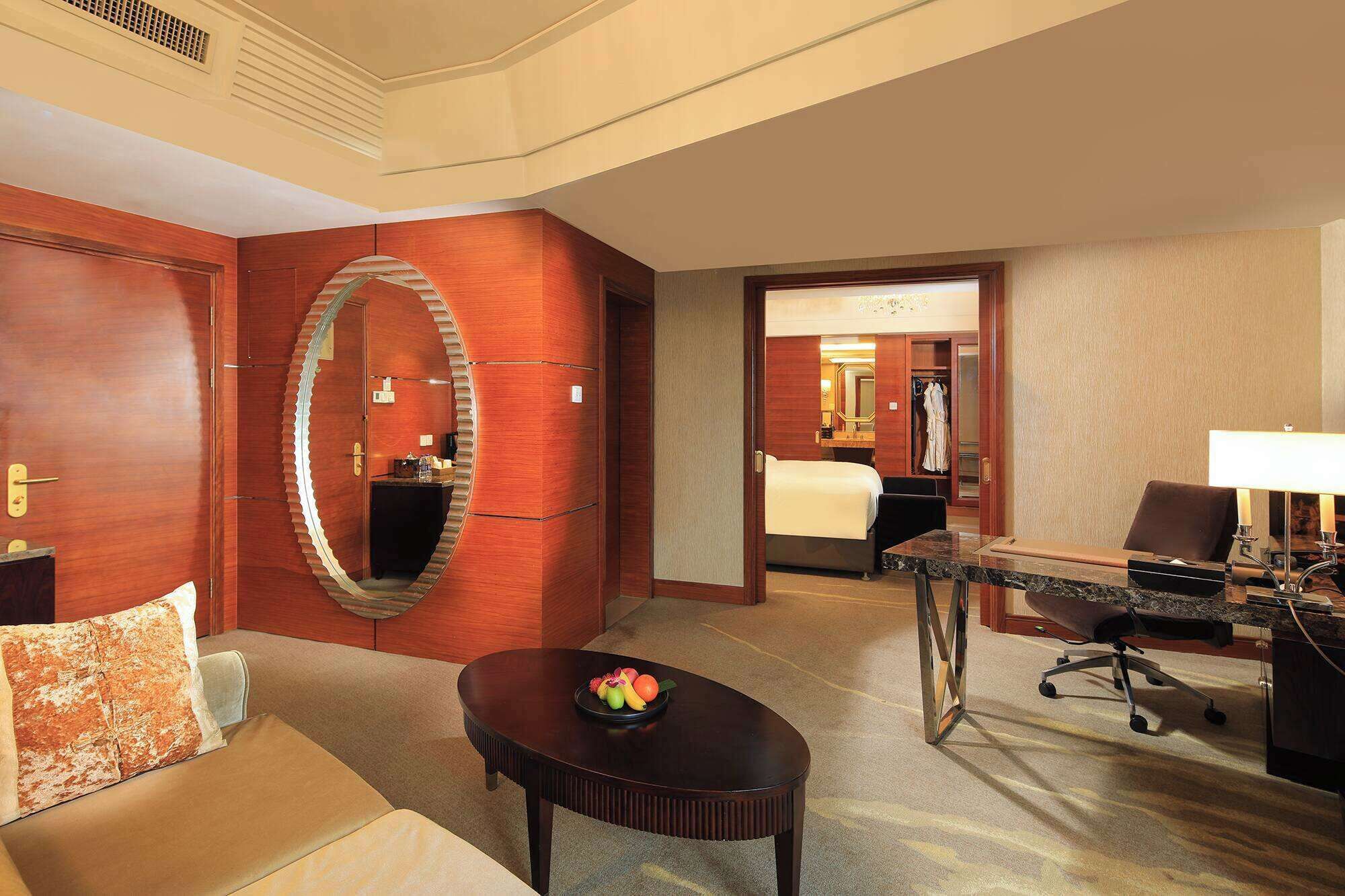 Une chambre d’hôtel avec un grand miroir, une table ronde sur laquelle sont disposés des fruits, un canapé, un bureau avec une lampe et un lit à l’arrière-plan.