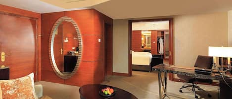 Une chambre d’hôtel avec un grand miroir, une table ronde sur laquelle sont disposés des fruits, un canapé, un bureau avec une lampe et un lit à l’arrière-plan.