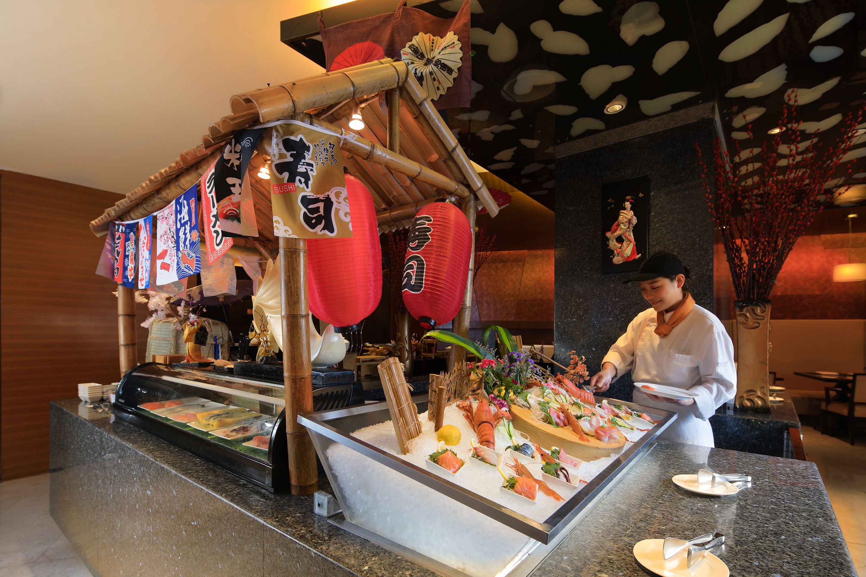 Un sushi bar con diversi tipi di pesce esposti su ghiaccio, uno chef che prepara il cibo e decorazioni tradizionali giapponesi.