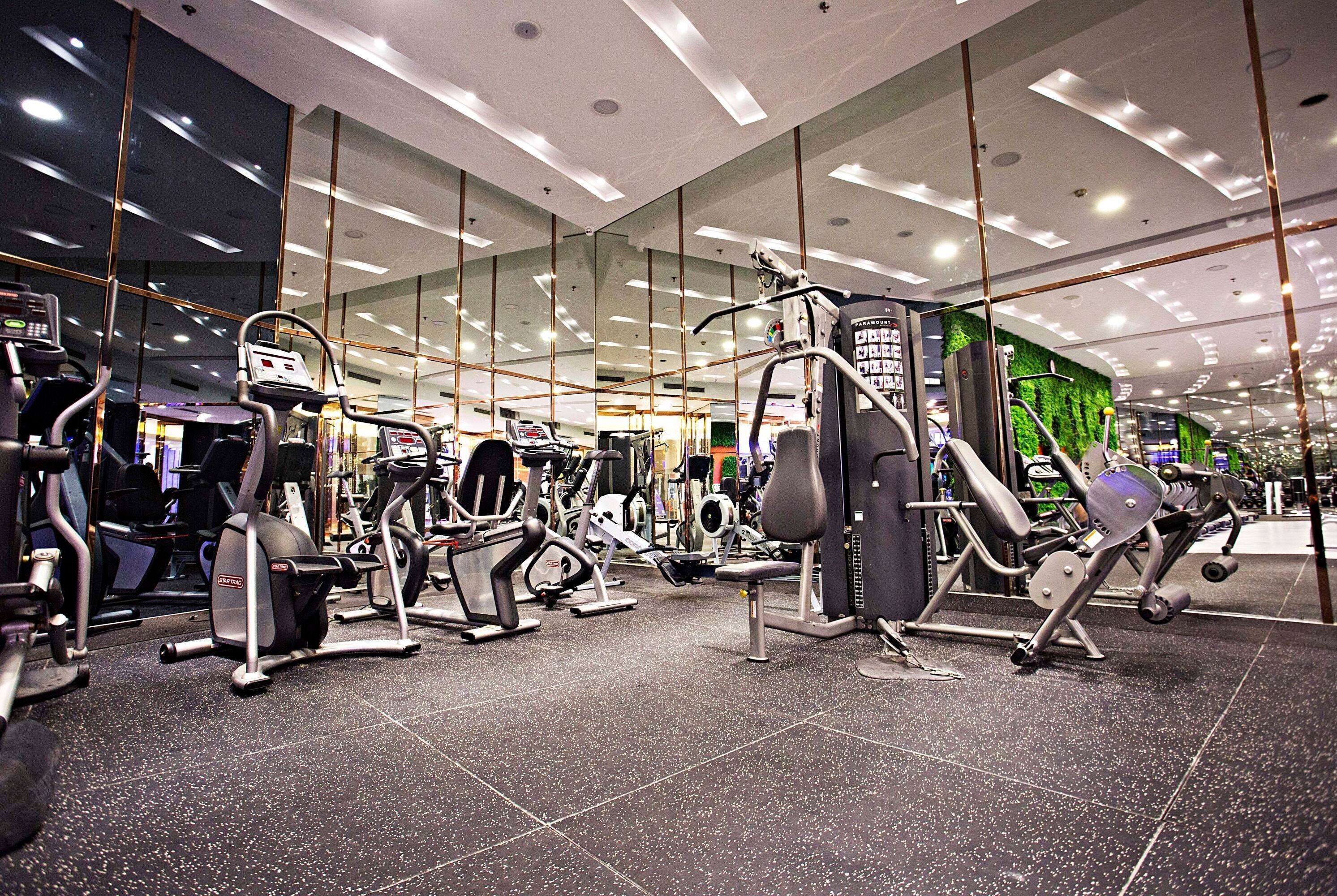 A modern gym with various exercise machines, large windows, and a green wall.