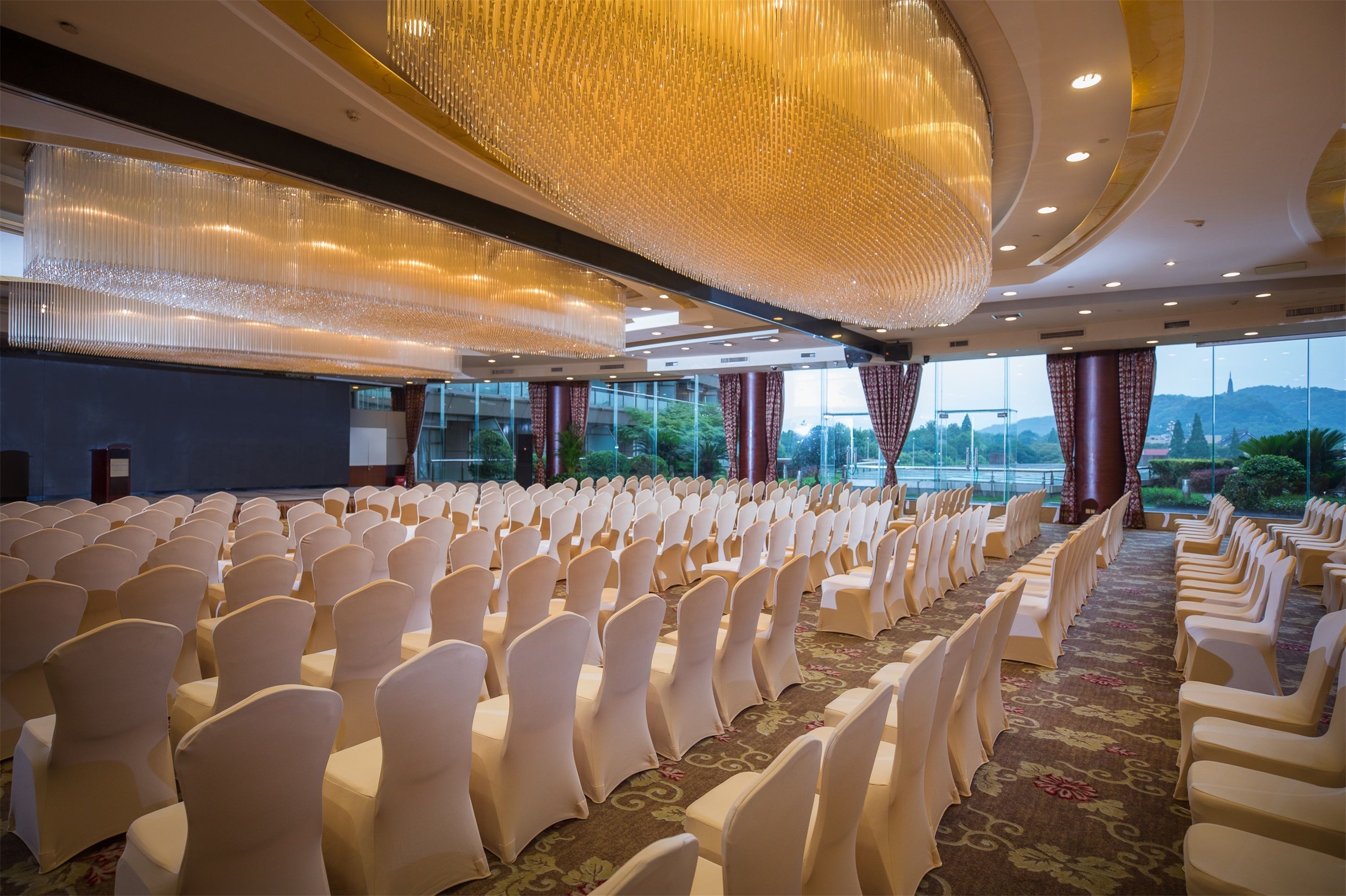 Une salle de conférence avec des rangées de chaises blanches, un grand lustre et une vue sur l’extérieur grâce à de grandes fenêtres.