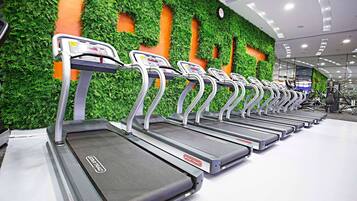 Varias cintas de correr en un gimnasio con un muro cubierto de follaje verde y un reloj en el techo.