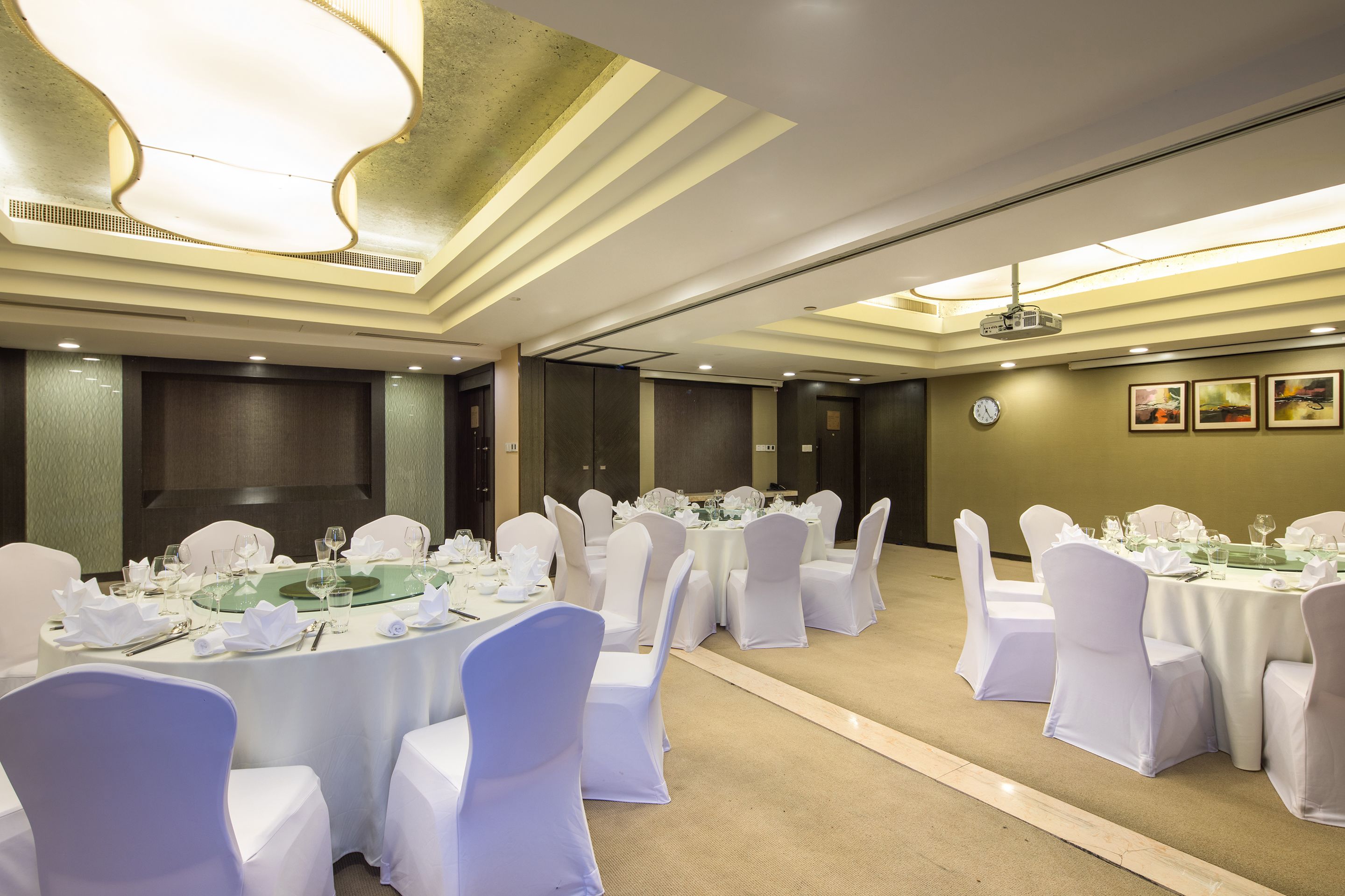 A banquet hall with round tables set for dining, white chairs, and a projector on the ceiling.