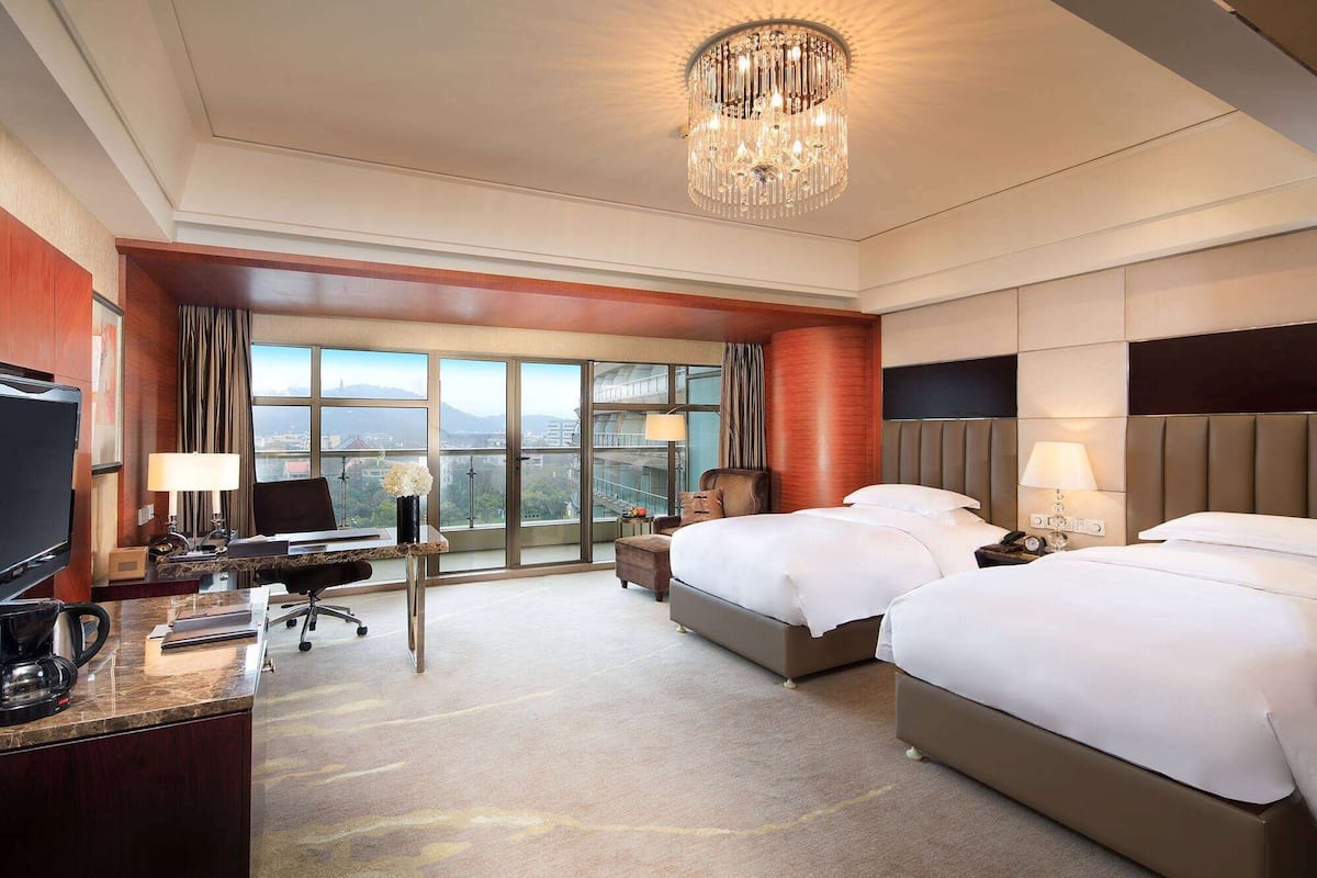 A hotel room with two beds, a desk with a computer, a TV, and a large window offering a city view.