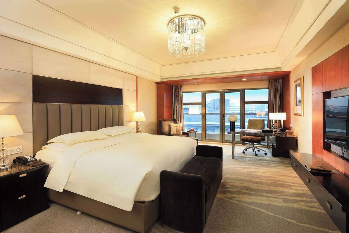 A hotel room with a large bed, a desk, a chair, a television, and a view of the city through the windows.